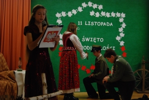 Uczniowie na scenie śpiewają pieśni. Koło sceny poczet sztandarowy, na scenie uczniowie przebrani w czarne stroje zaborców i  na biało za orła. Uczniowie składają znicze pod krzyżem.