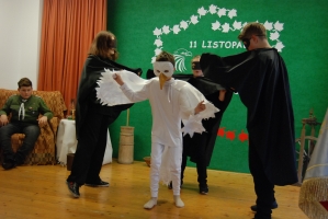 Uczniowie na scenie śpiewają pieśni. Koło sceny poczet sztandarowy, na scenie uczniowie przebrani w czarne stroje zaborców i  na biało za orła. Uczniowie składają znicze pod krzyżem.