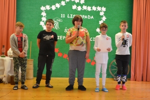 Uczniowie na scenie śpiewają pieśni. Koło sceny poczet sztandarowy, na scenie uczniowie przebrani w czarne stroje zaborców i  na biało za orła. Uczniowie składają znicze pod krzyżem.