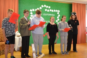 Uczniowie na scenie śpiewają pieśni. Koło sceny poczet sztandarowy, na scenie uczniowie przebrani w czarne stroje zaborców i  na biało za orła. Uczniowie składają znicze pod krzyżem.