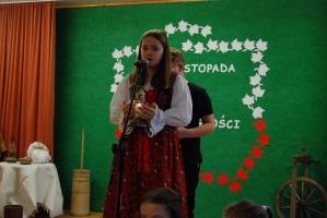 Uczniowie na scenie śpiewają pieśni. Koło sceny poczet sztandarowy, na scenie uczniowie przebrani w czarne stroje zaborców i  na biało za orła. Uczniowie składają znicze pod krzyżem.