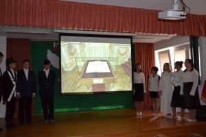 Uczniowie na scenie. Przebrani w stroje biało czarne i ułańskie z epoki. Tańczą, markują ruchy kopania, ogrzewania przy ognisku.