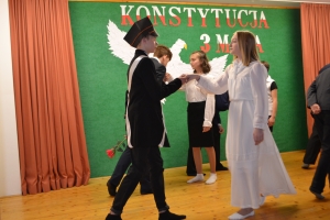 Uczniowie na scenie. Przebrani w stroje biało czarne i ułańskie z epoki. Tańczą, markują ruchy kopania, ogrzewania przy ognisku.