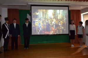 Uczniowie na scenie. Przebrani w stroje biało czarne i ułańskie z epoki. Tańczą, markują ruchy kopania, ogrzewania przy ognisku.