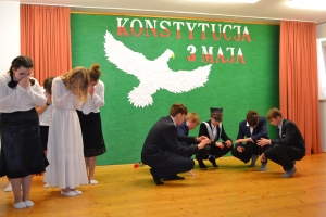 Uczniowie na scenie. Przebrani w stroje biało czarne i ułańskie z epoki. Tańczą, markują ruchy kopania, ogrzewania przy ognisku.