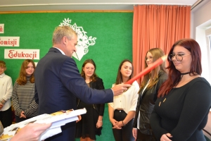 Uczniowie występują na scenie w galowych strojach, dyrektor pasuje uczniów długim ołówkiem