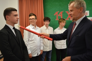 Uczniowie występują na scenie w galowych strojach, dyrektor pasuje uczniów długim ołówkiem