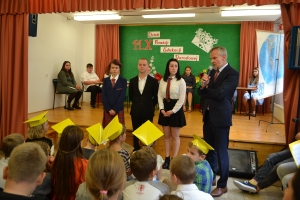 Uczniowie występują na scenie w galowych strojach, dyrektor pasuje uczniów długim ołówkiem