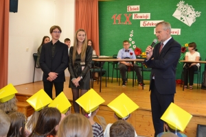 Uczniowie występują na scenie w galowych strojach, dyrektor pasuje uczniów długim ołówkiem