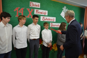 Uczniowie występują na scenie w galowych strojach, dyrektor pasuje uczniów długim ołówkiem