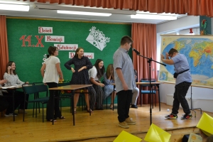 Uczniowie występują na scenie w galowych strojach, dyrektor pasuje uczniów długim ołówkiem