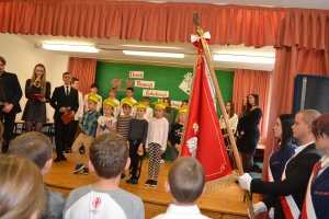 Uczniowie występują na scenie w galowych strojach, dyrektor pasuje uczniów długim ołówkiem