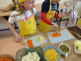 dzieci ubrane w fartuszki i czapki kucharskie, kroją warzywa